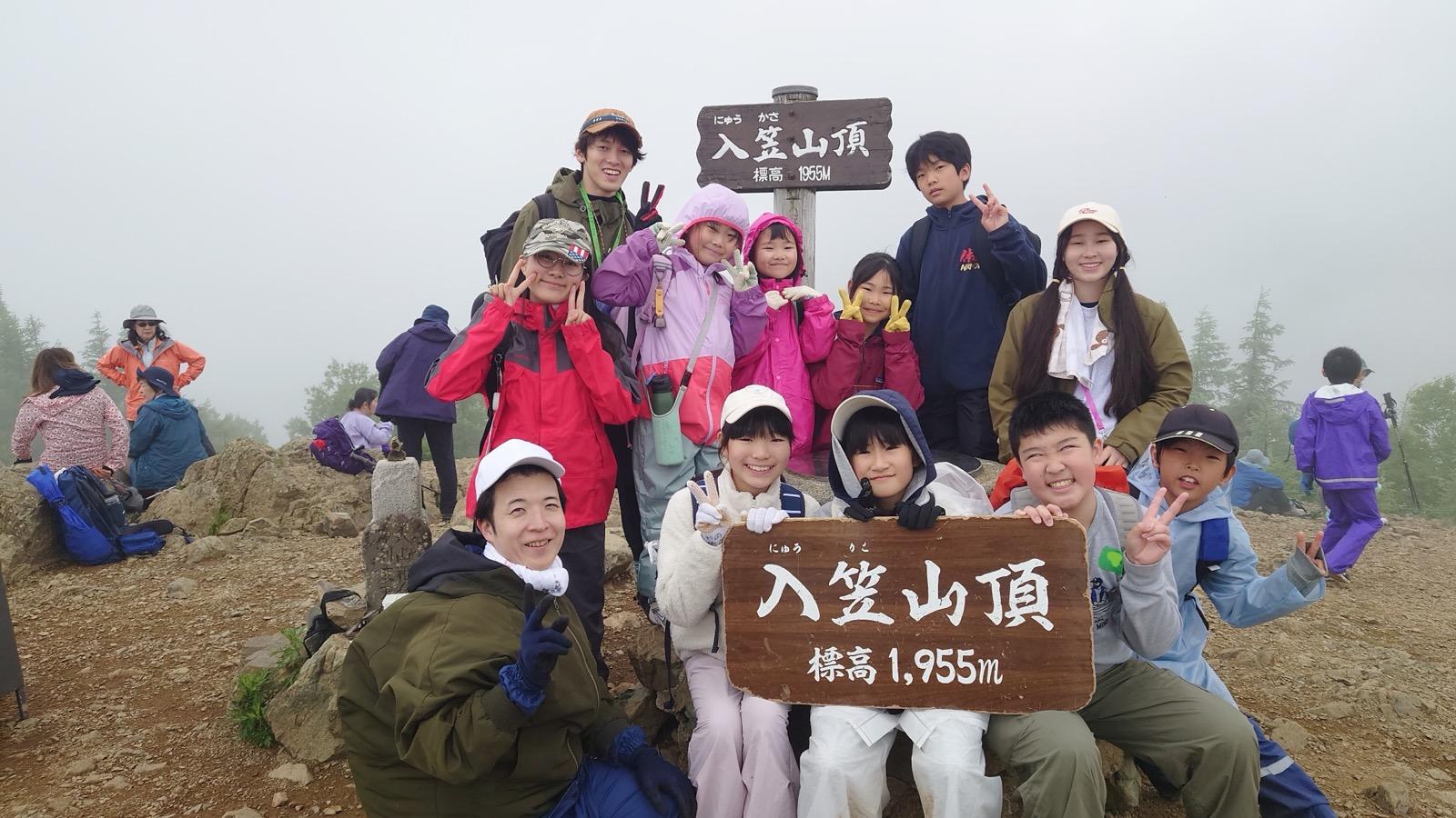 霧に包まれた入笠山の山頂で、「入笠山頂 標高1,955m」の看板を子どもたちが持ち、リーダーと一緒に記念撮影をしている