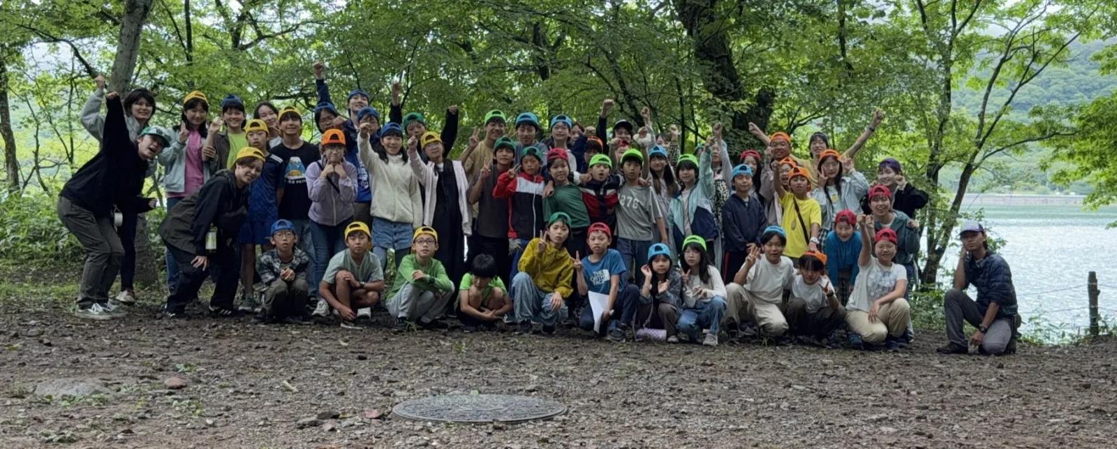 大きな木々に囲まれた広場で、参加者とスタッフ全員が湖を背に集まって記念撮影をしている
